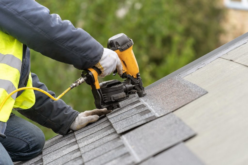 Unrecognizable roofer worker in uniform work wear using air or pneumatic nail gun and installing asphalt or bitumen tile on top of the roof under construction house