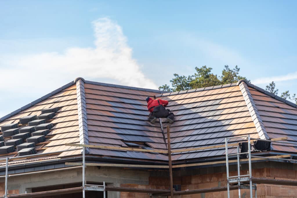unfinished building, roofer worker installing grey metal tile on top of roof outdoors on blue sky background