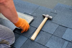 Contractor hands installing bitumen roof shingles using hammer and nails.